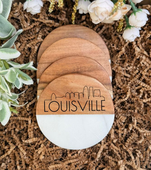 Four round wooden coasters stacked on top of each other, with the front coaster featuring a white lower half and an engraved black Louisville skyline and text. The coasters are photographed on brown shredded paper with decorative greenery and white flowers around them.