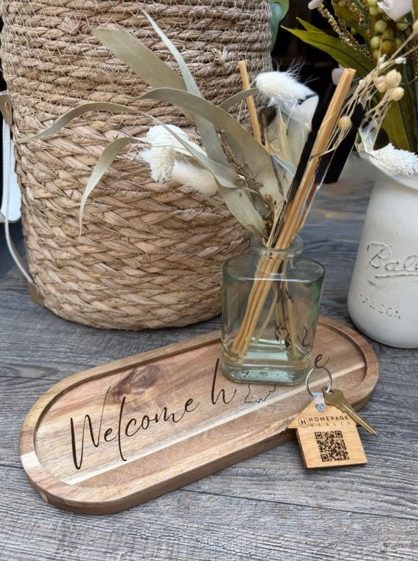 A wooden welcome sign tray engraved with 'Welcome home' in script, styled with a small clear glass reed diffuser and a hanging wooden brand tag on a gray wood surface. A woven basket and a white Ball jar with dried flowers are in the background.
